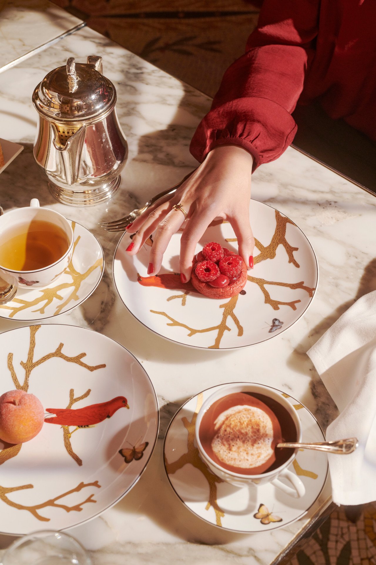 Alguns pormenores da hora do chá com uma mão a tirar uma bolacha de framboesa de Cédric Grolet ao lado de bebidas quentes e um pêssego, no Le Meurice, Paris  